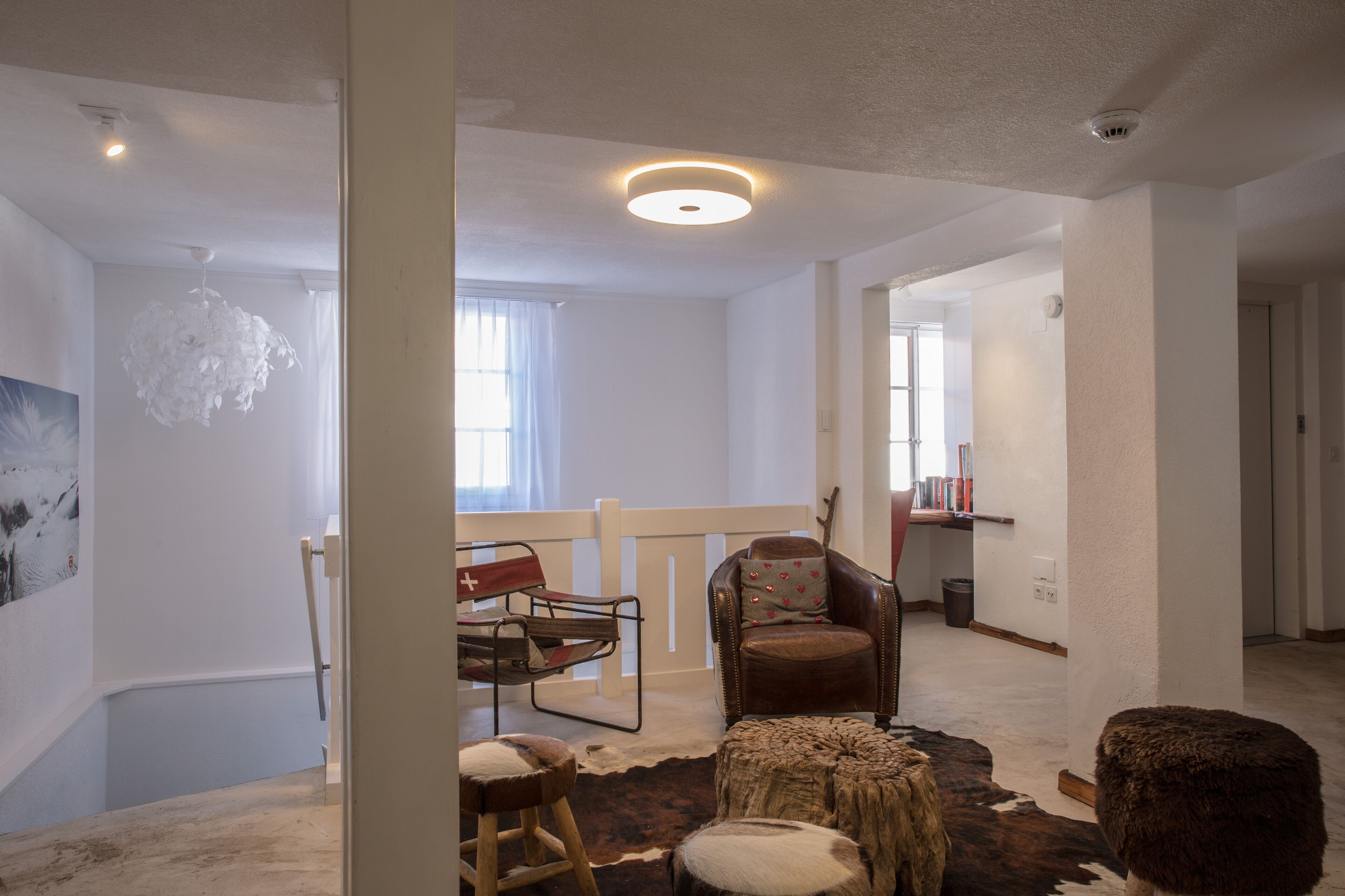 Una habitación amplia con un gran ventanal, una silla de madera, un sillón de cuero, una mesita redonda y una lámpara de techo blanca.