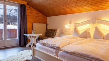Ein gemütliches Schlafzimmer mit Holzbalkendecke, einem Bett mit weißer Bettwäsche, einem kleinen Tisch mit Lampe und Blick auf schneebedeckte Berge durch eine Schiebetür.