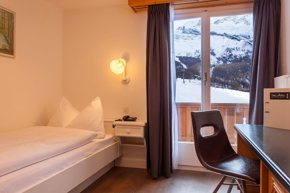 A hotel room with a bed, a desk, a chair, a nightstand, a lamp, and a view of a snowy mountain landscape.