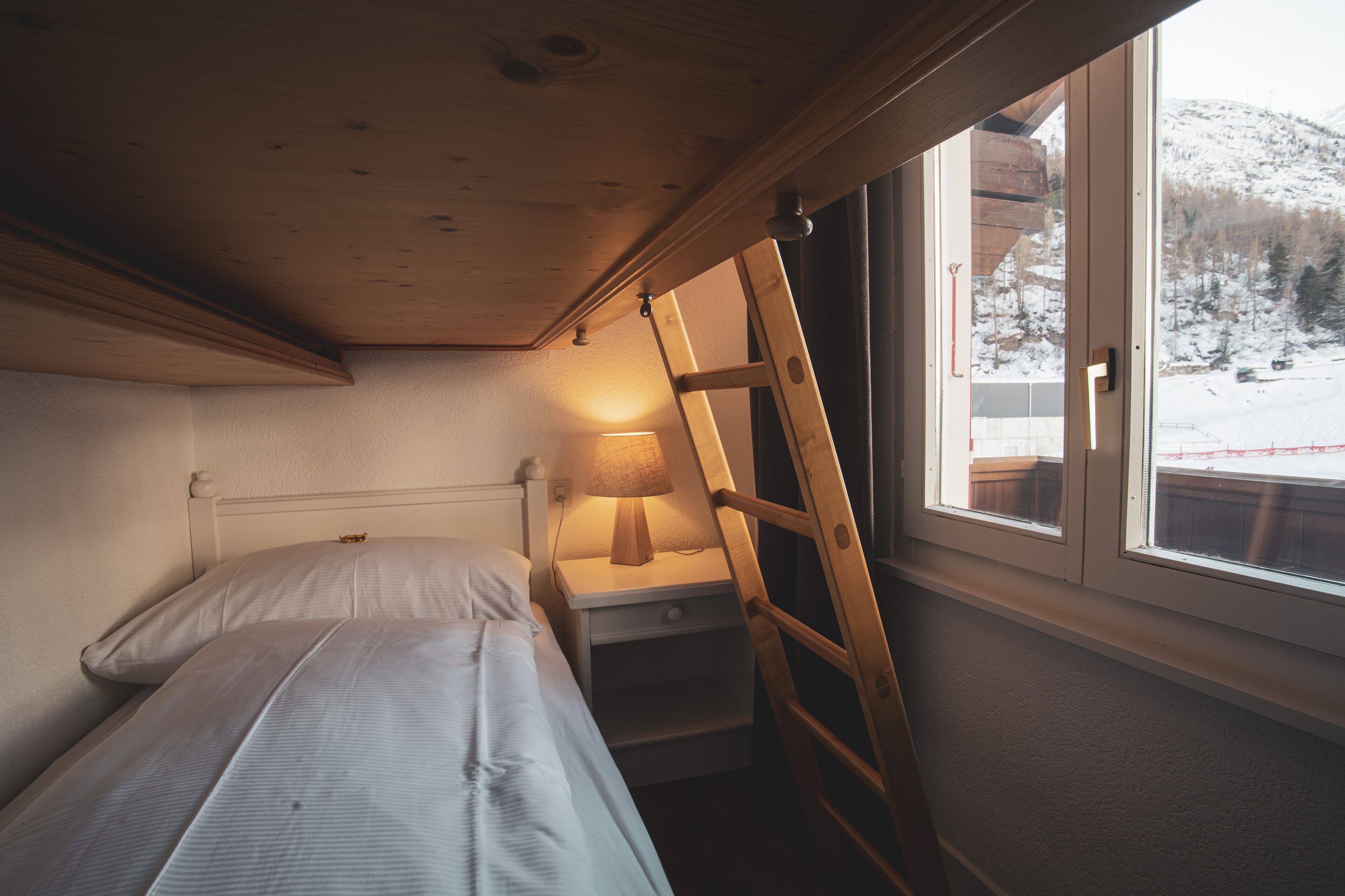 A small, cozy room with a bed, a nightstand, a lamp, and a window with a view of a snowy landscape.