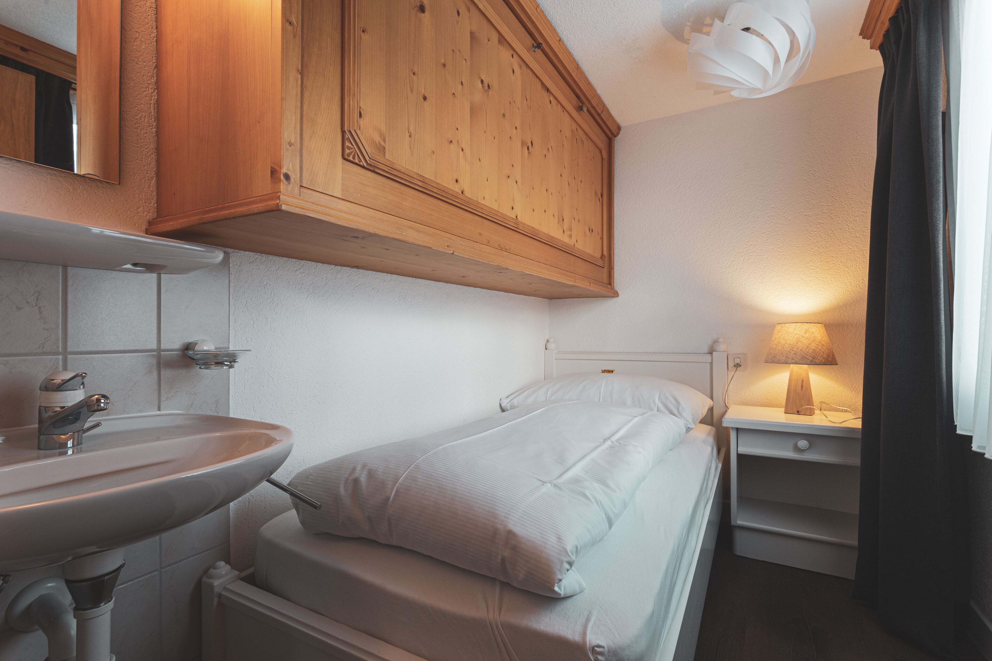 A small, neatly organized bedroom with a single bed, a bedside table with a lamp, a wooden cabinet above the bed, and a bathroom with a sink and mirror.