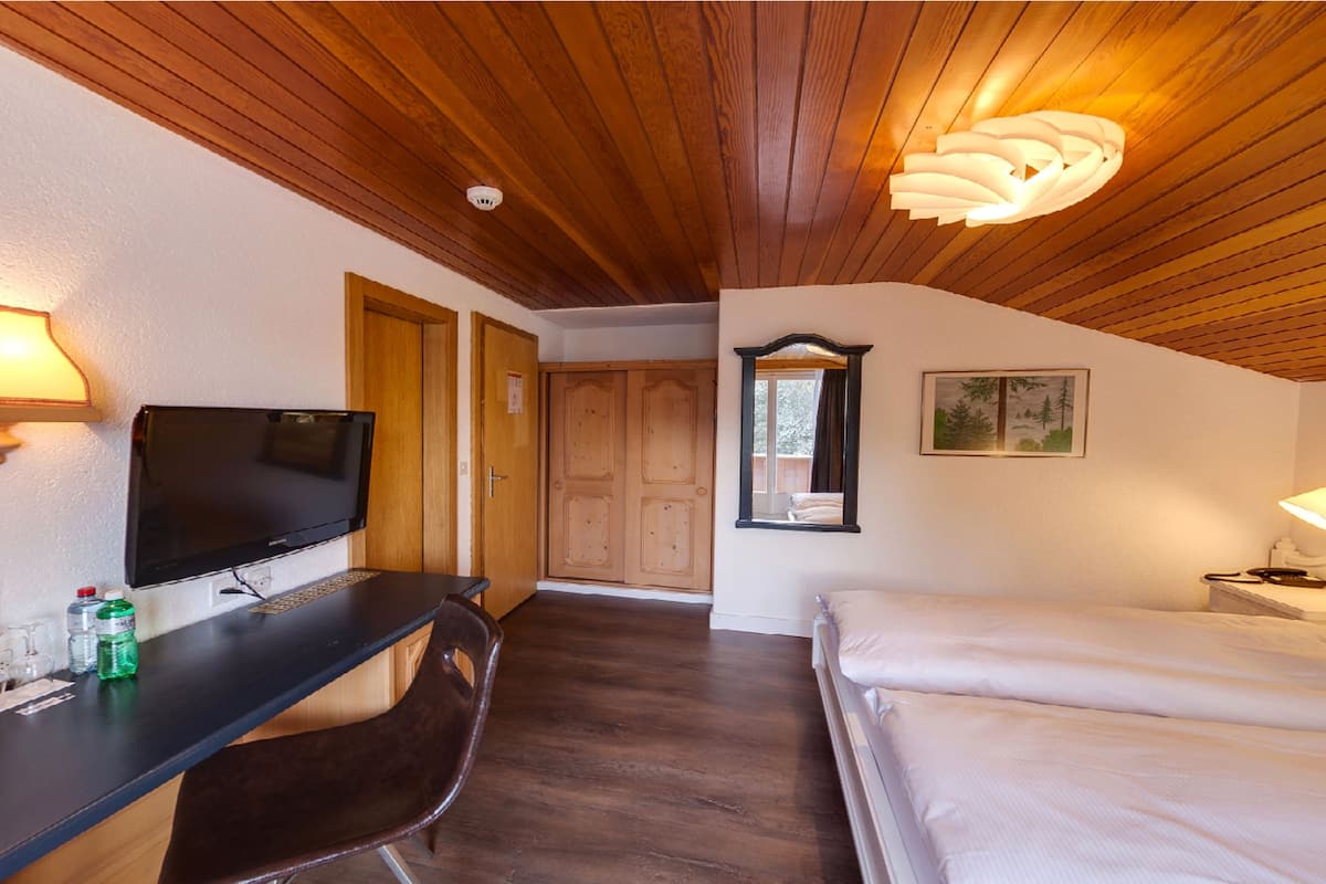A room with a wooden ceiling, a flat-screen TV, a desk with a chair, a bed, and a window with a view of trees.
