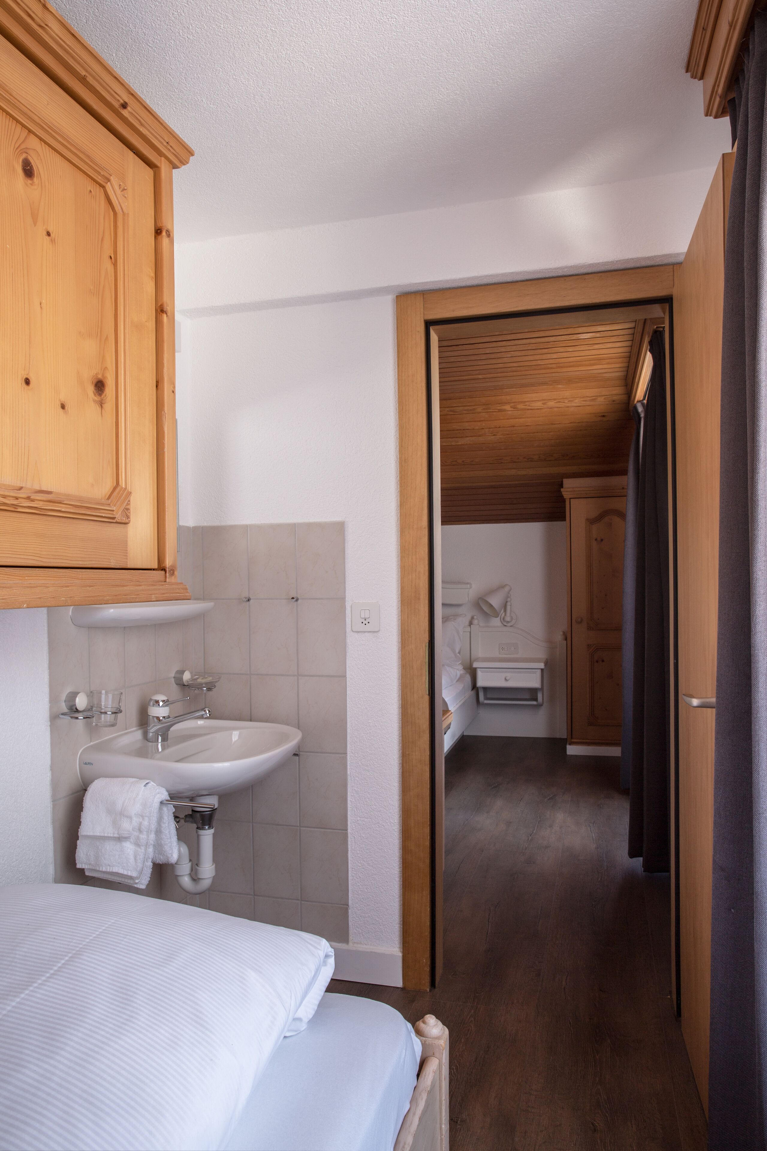 Un pasillo estrecho con una puerta de madera que da a un baño. Se ve una cama con ropa blanca a la izquierda.