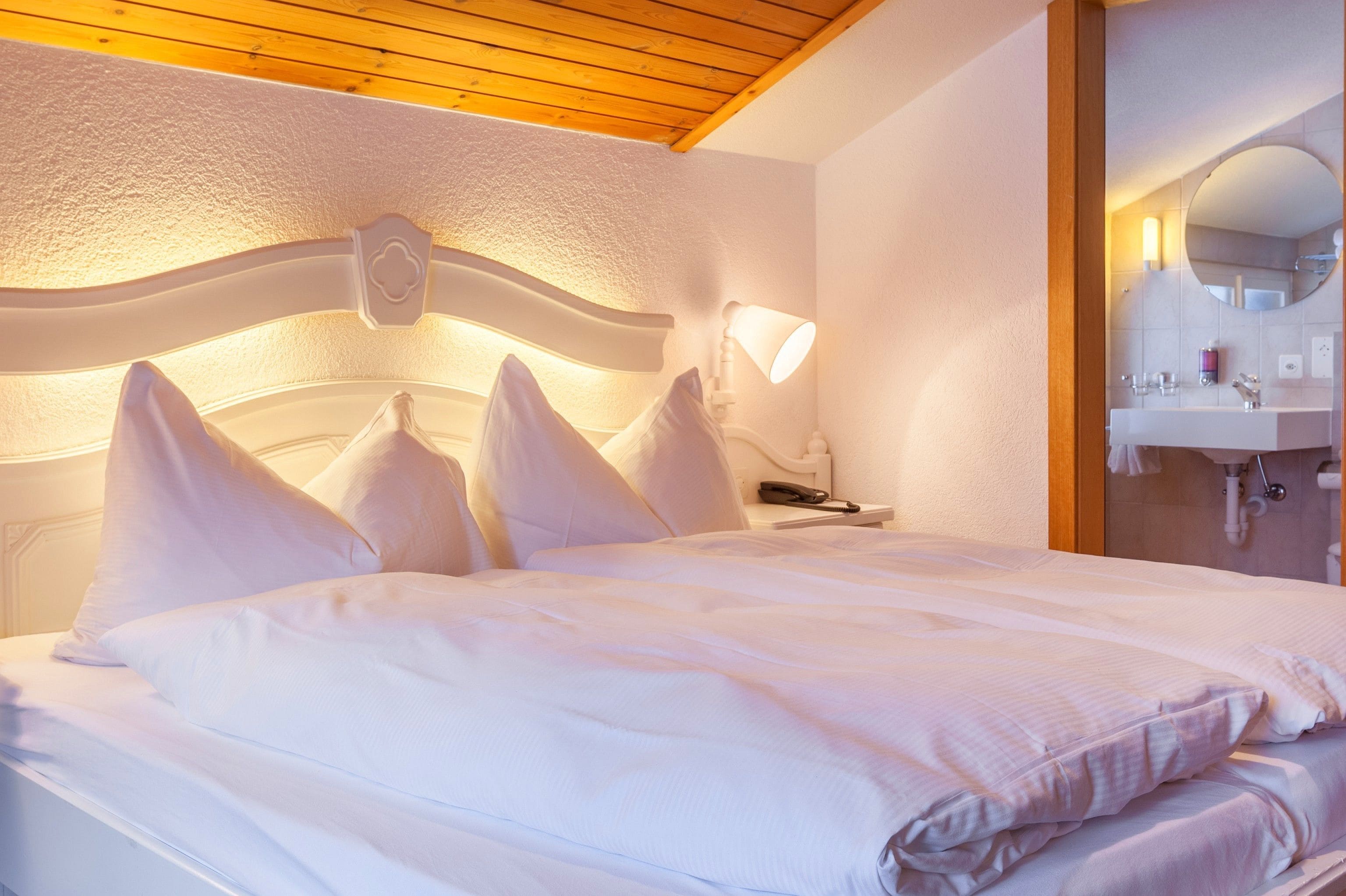 Ett hotellrum med en säng, kuddar, ett sängbord med en lampa, en spegel och ett badrum med handfat och hygienartiklar.