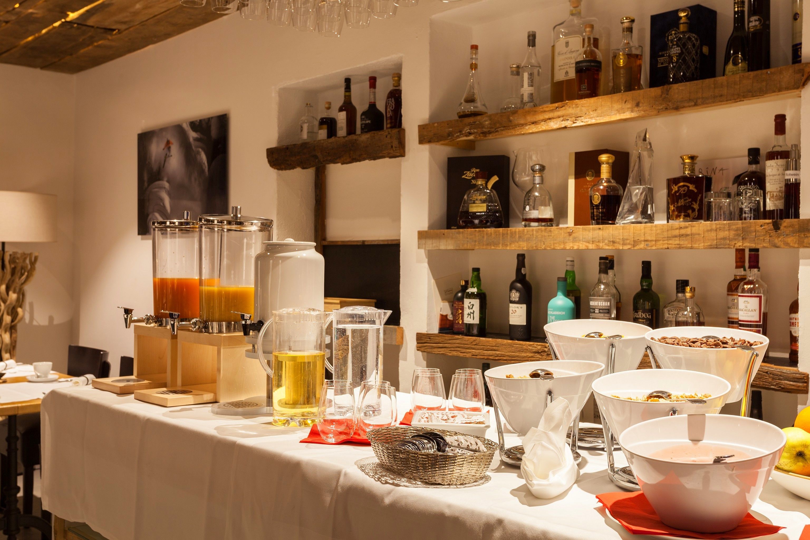 Un bufé de desayuno con diversas opciones de comida y bebidas, incluyendo un bar de zumos y una selección de botellas de alcohol en estanterías.