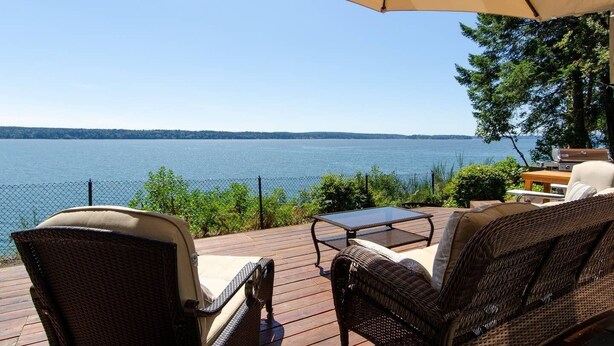 A deck with wicker furniture, a glass table, and an umbrella overlooking a lake. - Island house located on Stretch Island close to Fair Harbor (Grapeview)