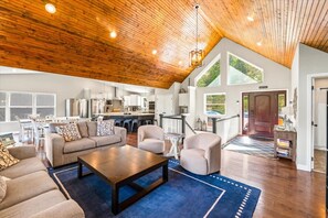 A spacious living area with a high ceiling, a kitchen in the background, and a view of the outdoors.