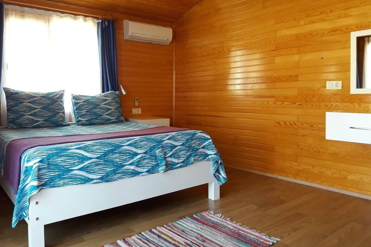 A wooden-paneled room with a bed, a window with curtains, an air conditioning unit, and a small table.