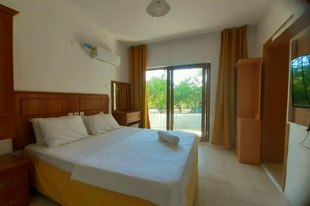 A hotel room with a large bed, wooden headboard, a nightstand, a wardrobe, and a balcony with curtains.