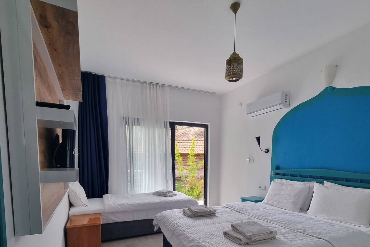 A modern hotel room with two beds, a wooden sideboard, a small wooden stool, and a view of greenery outside.