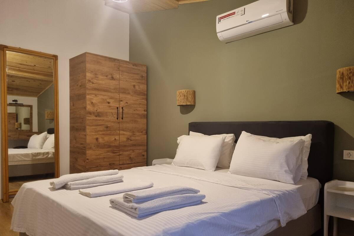 A neatly made bed with white linens and towels, a wooden wardrobe, a wall-mounted air conditioner, and a mirror reflecting part of the room.