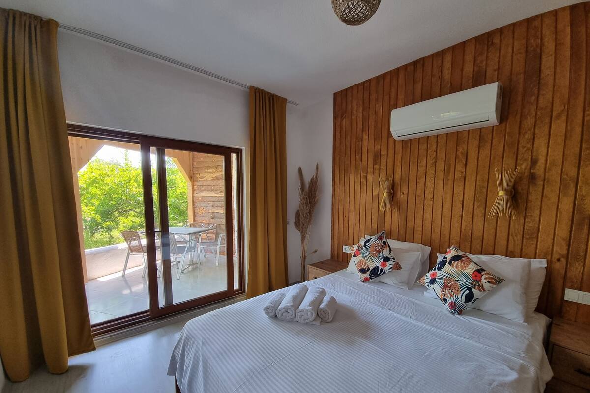 A bedroom with a large bed, a wooden wall, an air conditioning unit, and a view of an outdoor patio with a table and chairs.
