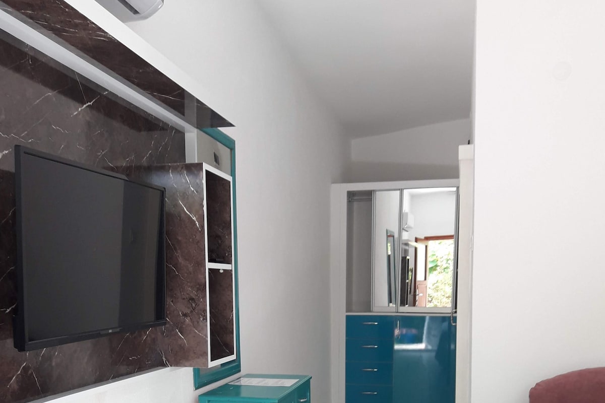 Habitación de hotel con televisión montada en una pared de mármol, un escritorio verde azulado, un sofá rojo y un baño con armarios azules.