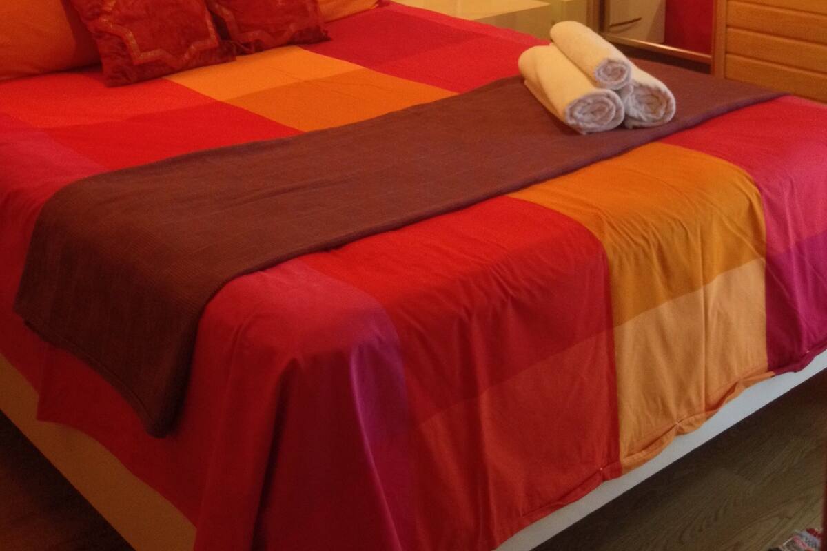 A room with a wooden wall, a bed with a red and orange bedspread, and a mirror reflecting the bed and a red pillow.