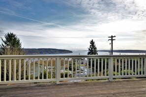 A wooden deck with a white railing overlooking a residential area with houses and a body of water in the distance.