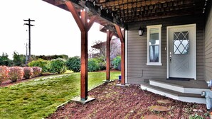 Eine überdachte Veranda mit Holzbalken, ein graues Haus mit einer weißen Tür und ein angelegter Garten mit rotem Rindenmulch und trittfesten Steinen.