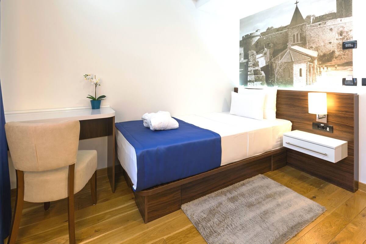 A hotel room with a bed, a desk, a chair, a small potted plant, and a wall-mounted nightstand with a lamp.