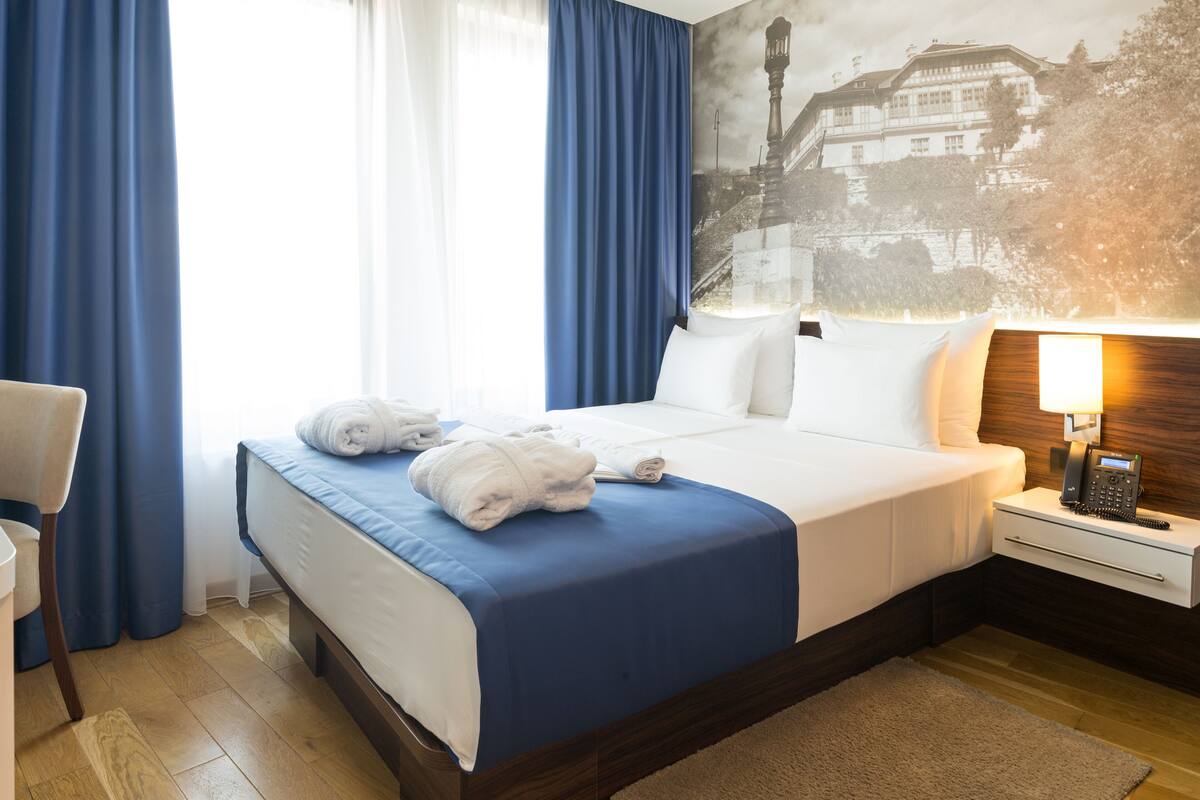 A hotel room with a neatly made bed, a bedside table with a lamp, and a large window with curtains.