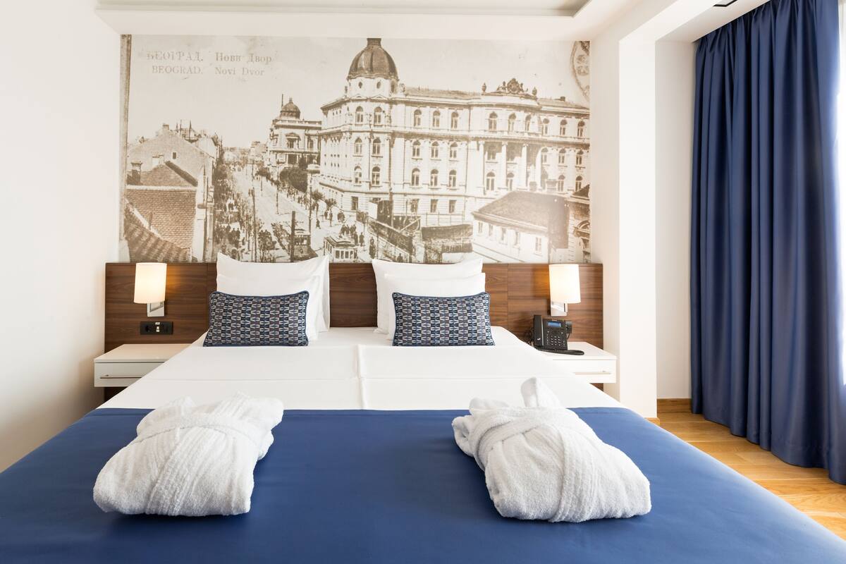 A hotel room with a large bed, two pillows, a nightstand, a lamp, and a wall-mounted picture.