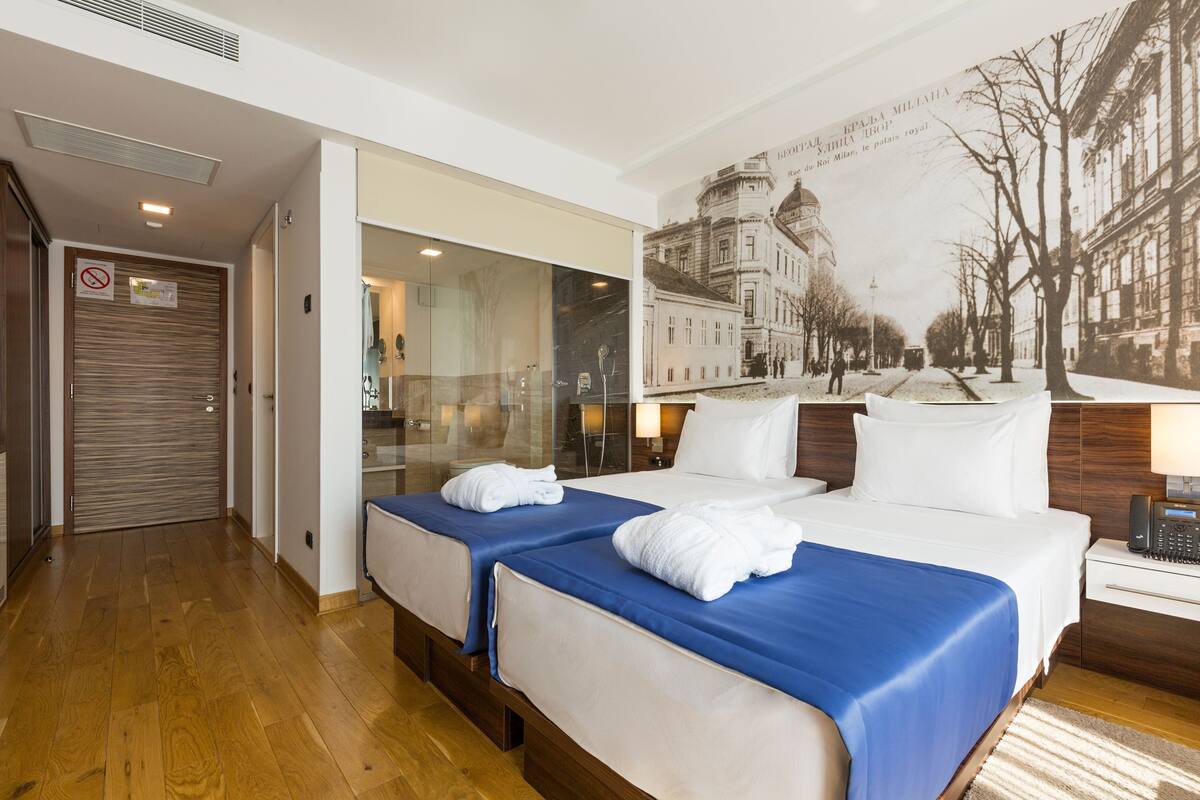 A hotel room with two beds, a large mirror, a painting of a street scene, and a telephone on the nightstand.