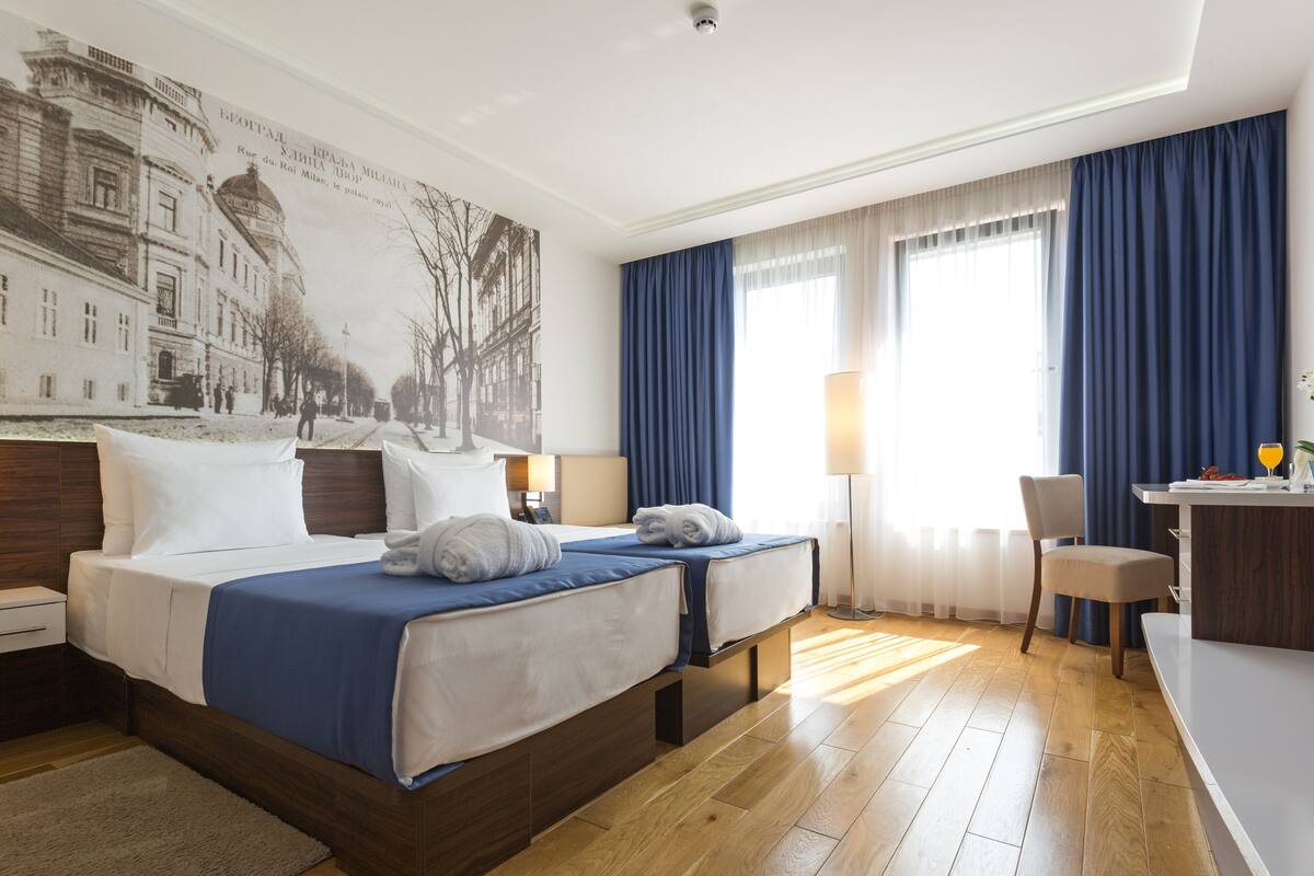 A hotel room with two beds, a desk, a chair, and a large window with curtains.