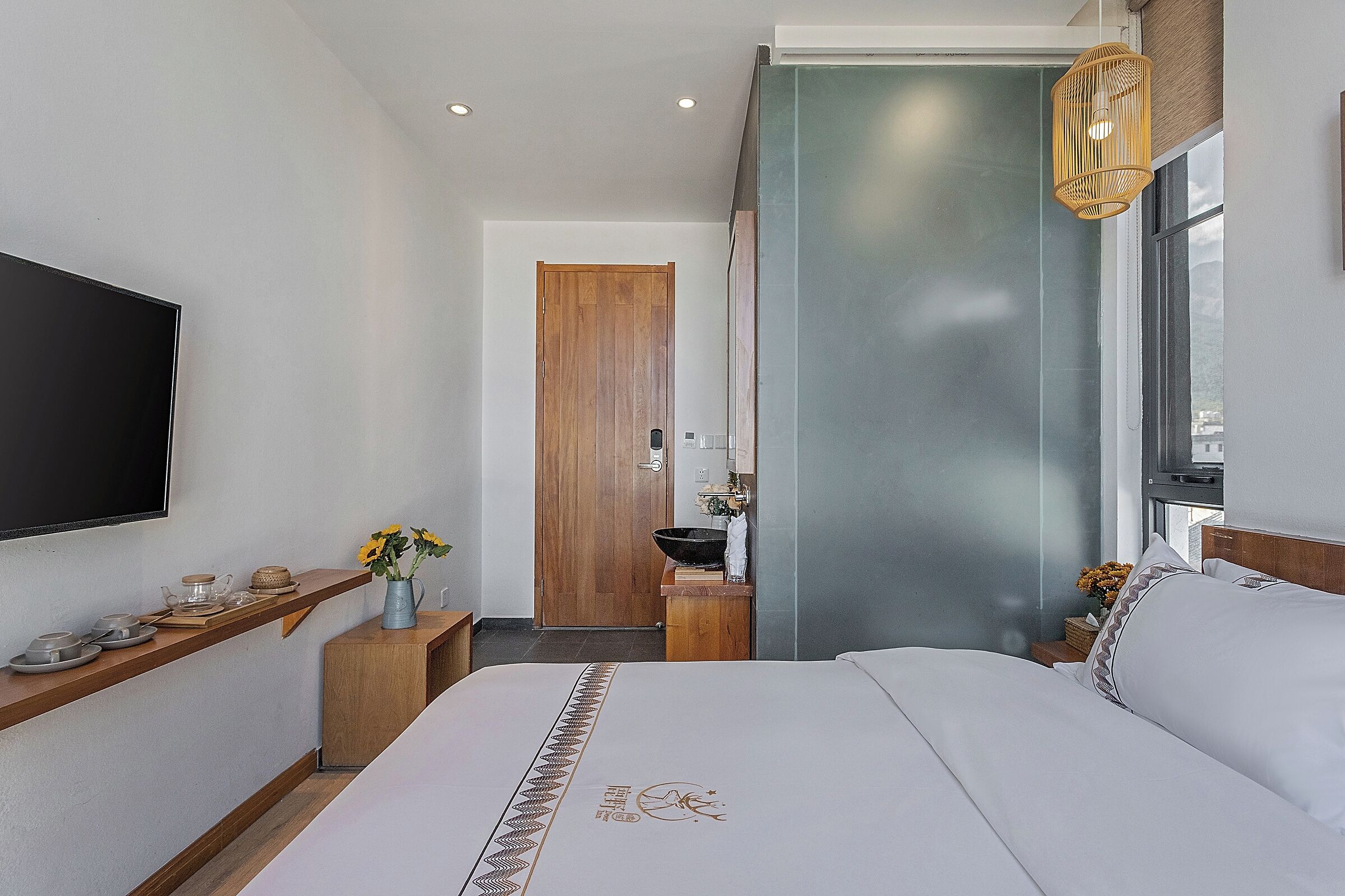 A modern hotel room with a bed, a mounted TV, a wooden door, a small bathroom, and a window.