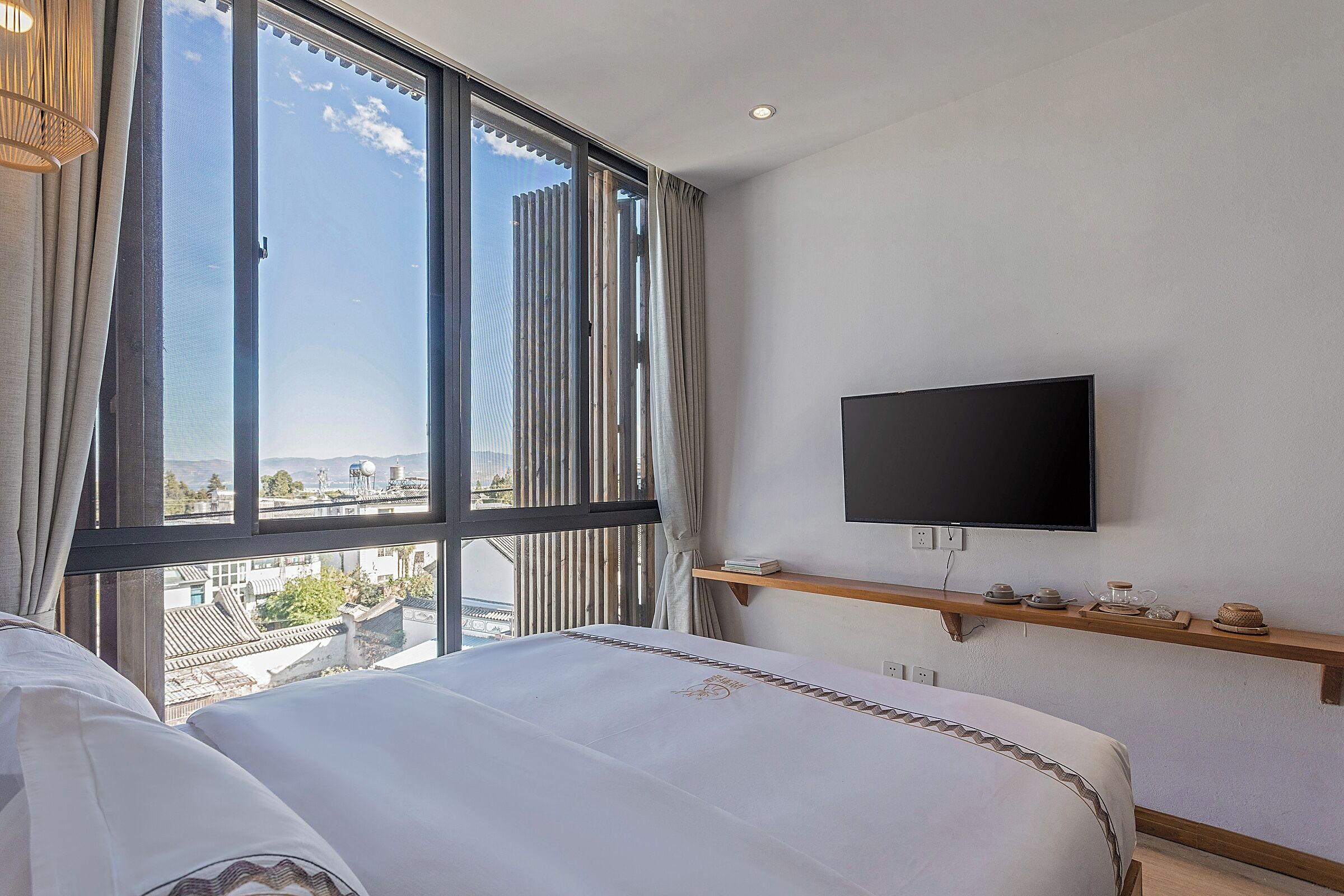 A hotel room with a large window, a bed, a mounted TV, and a shelf with items.