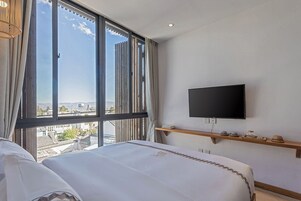 Una habitación de hotel con un ventanal grande, una cama, un televisor montado y una estantería con objetos.