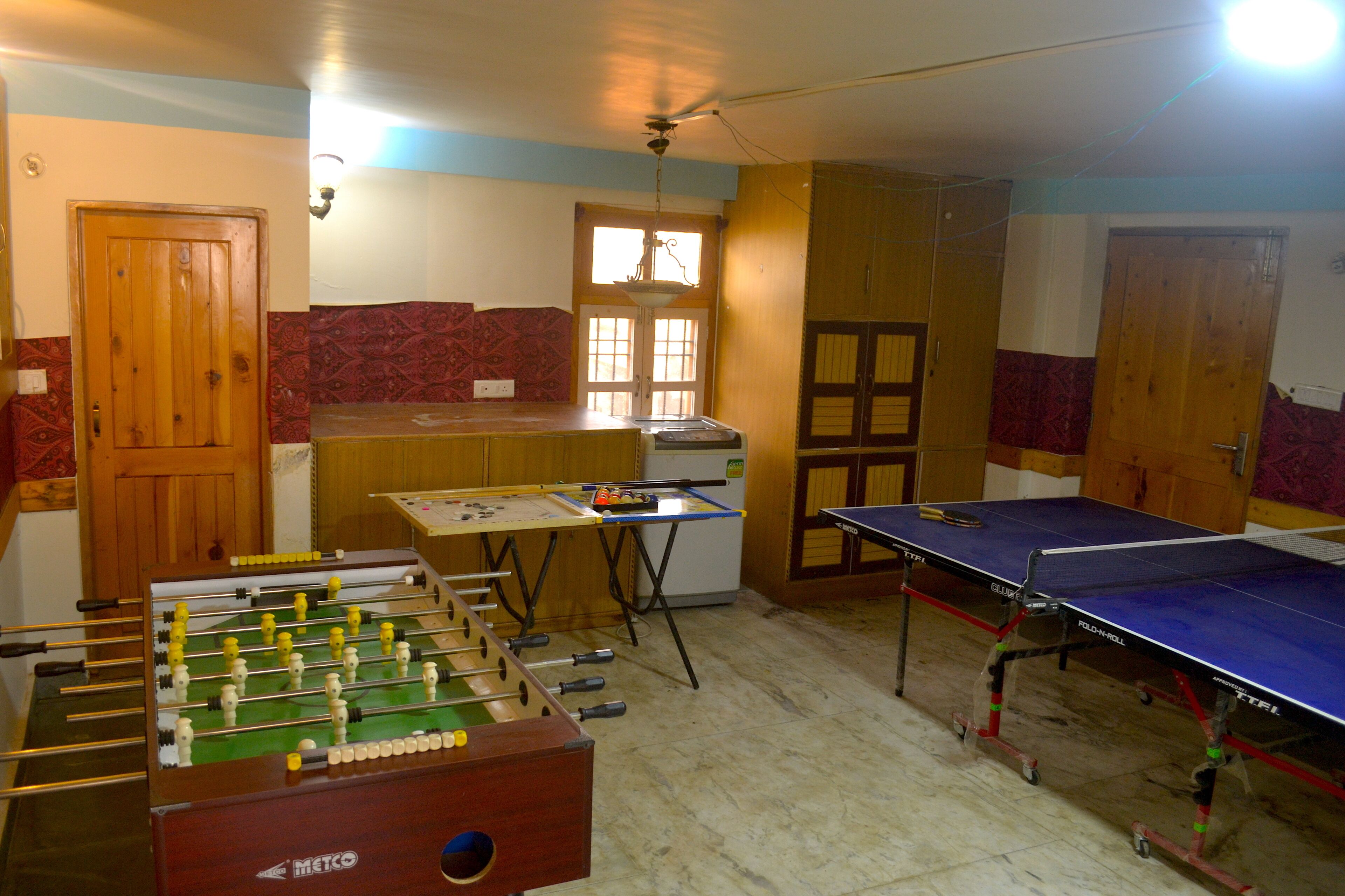 A room with a foosball table, a ping pong table, and a refrigerator.