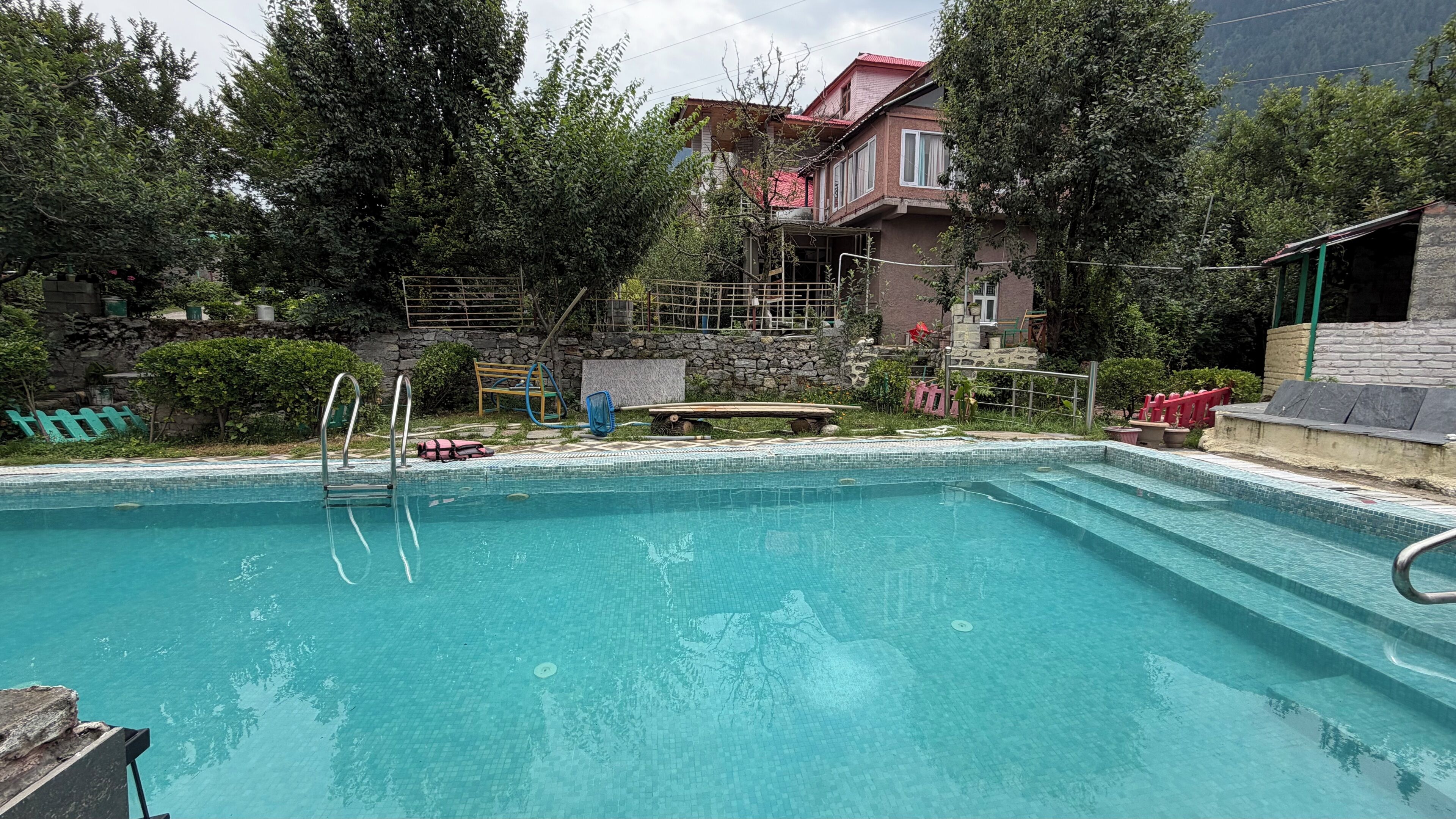 A swimming pool with steps leading into it, surrounded by greenery and a stone wall.