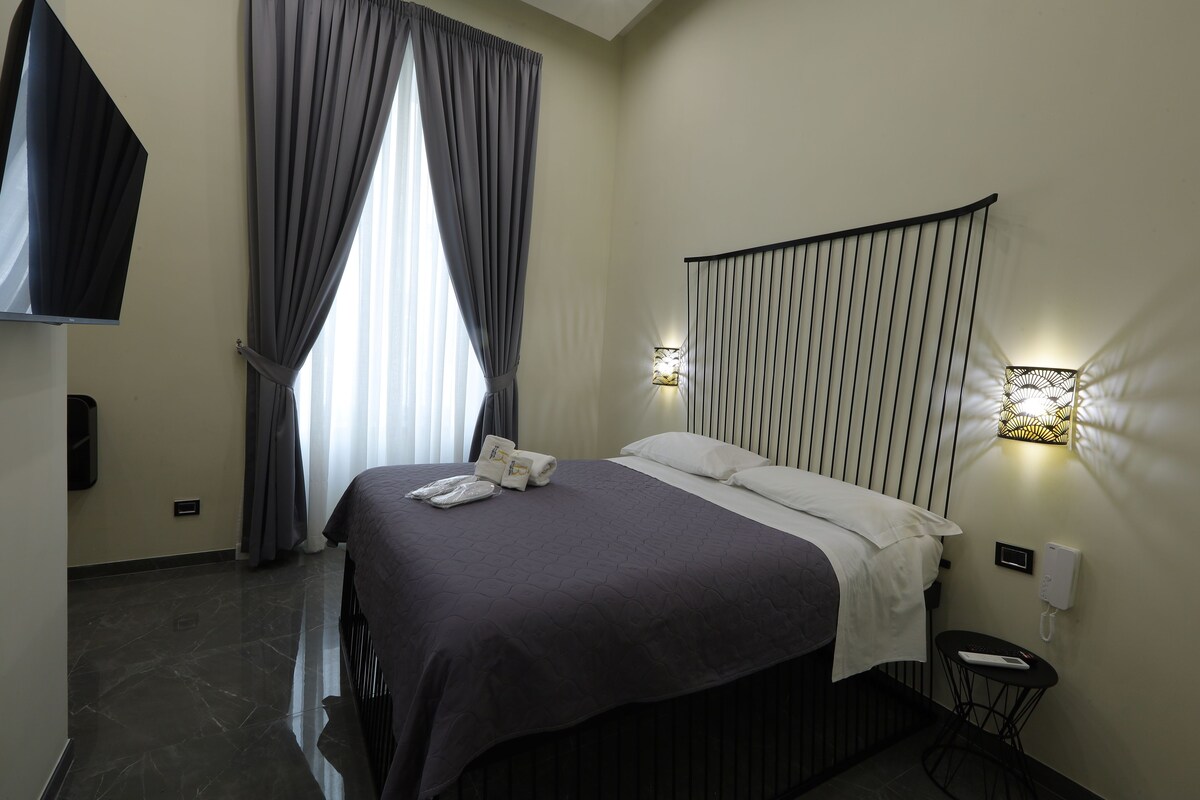 Una habitación de hotel con cama, cortinas, televisor, luces empotradas en la pared y una mesita de noche.