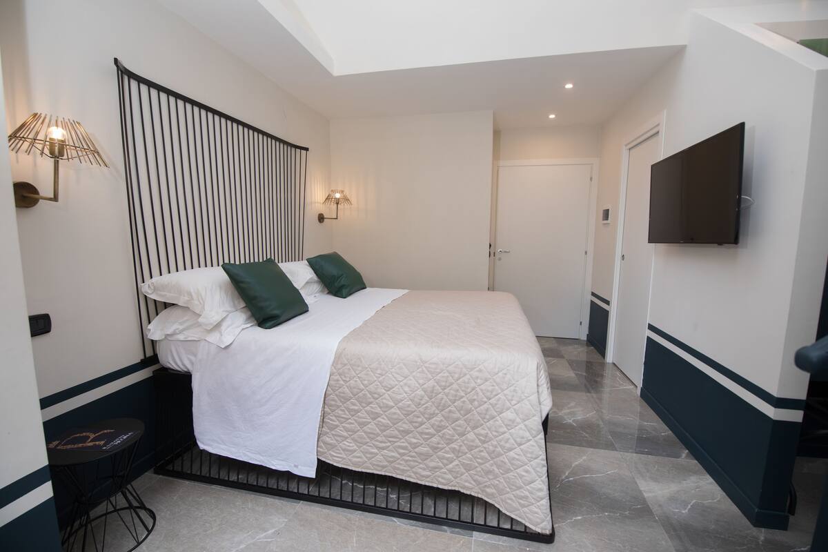 Une chambre à coucher bien rangée, avec un lit, des lampes de chevet, un téléviseur à écran plat et une porte donnant sur une autre pièce.