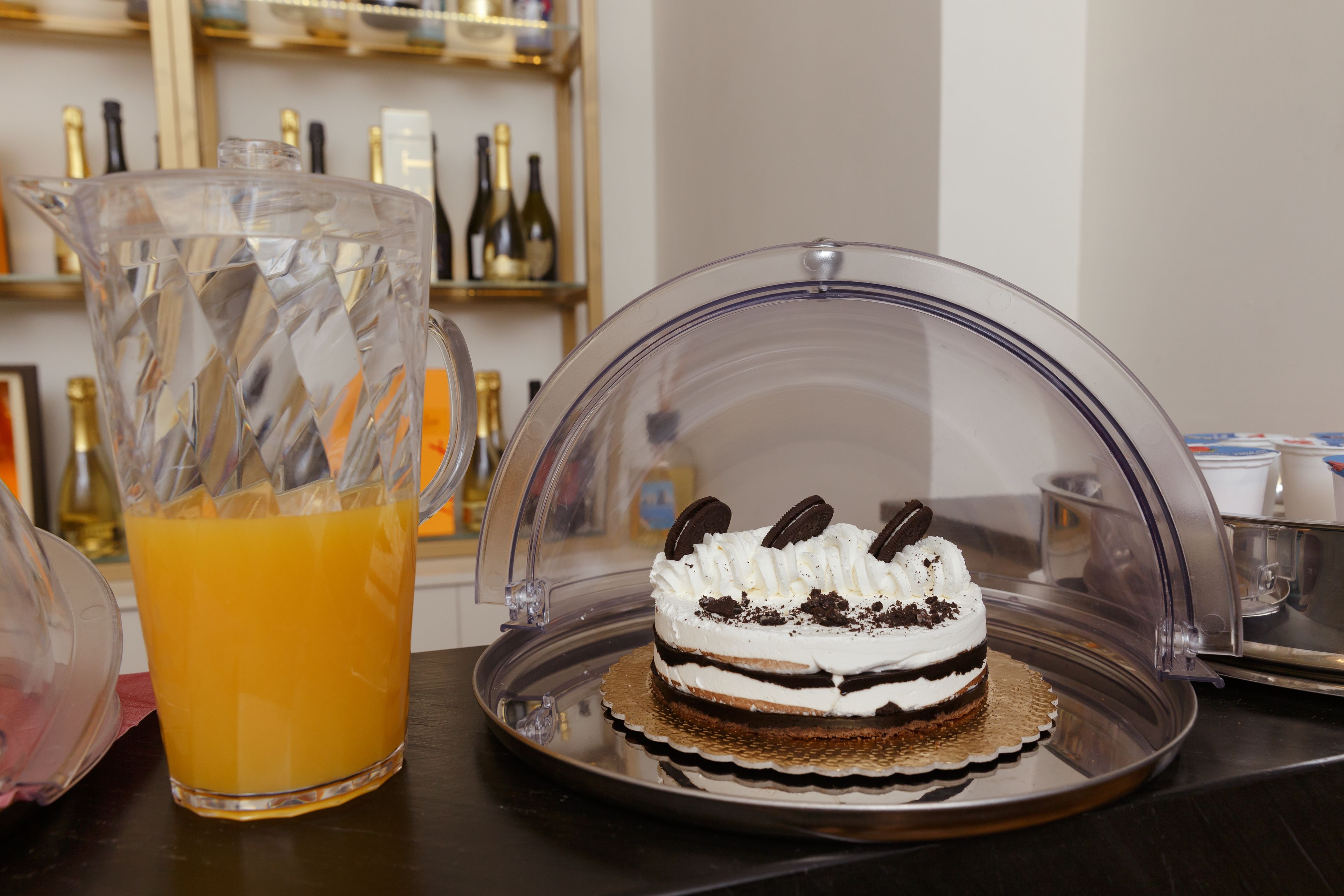 A dessert with a clear dome cover, a pitcher of orange juice, and bottles in the background.