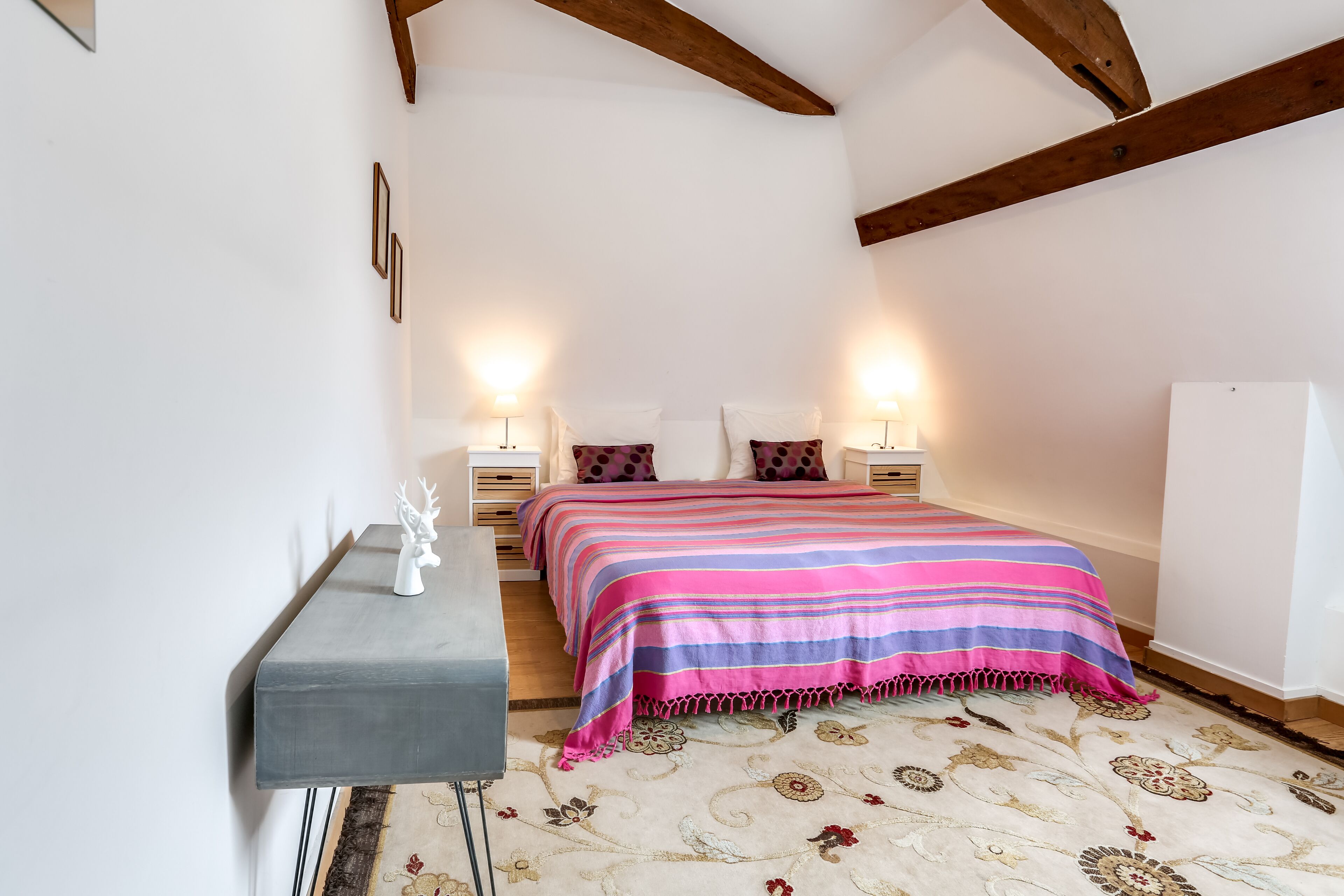 A bedroom with a bed, bedside tables, lamps, a nightstand, a decorative deer figurine, and a patterned rug.