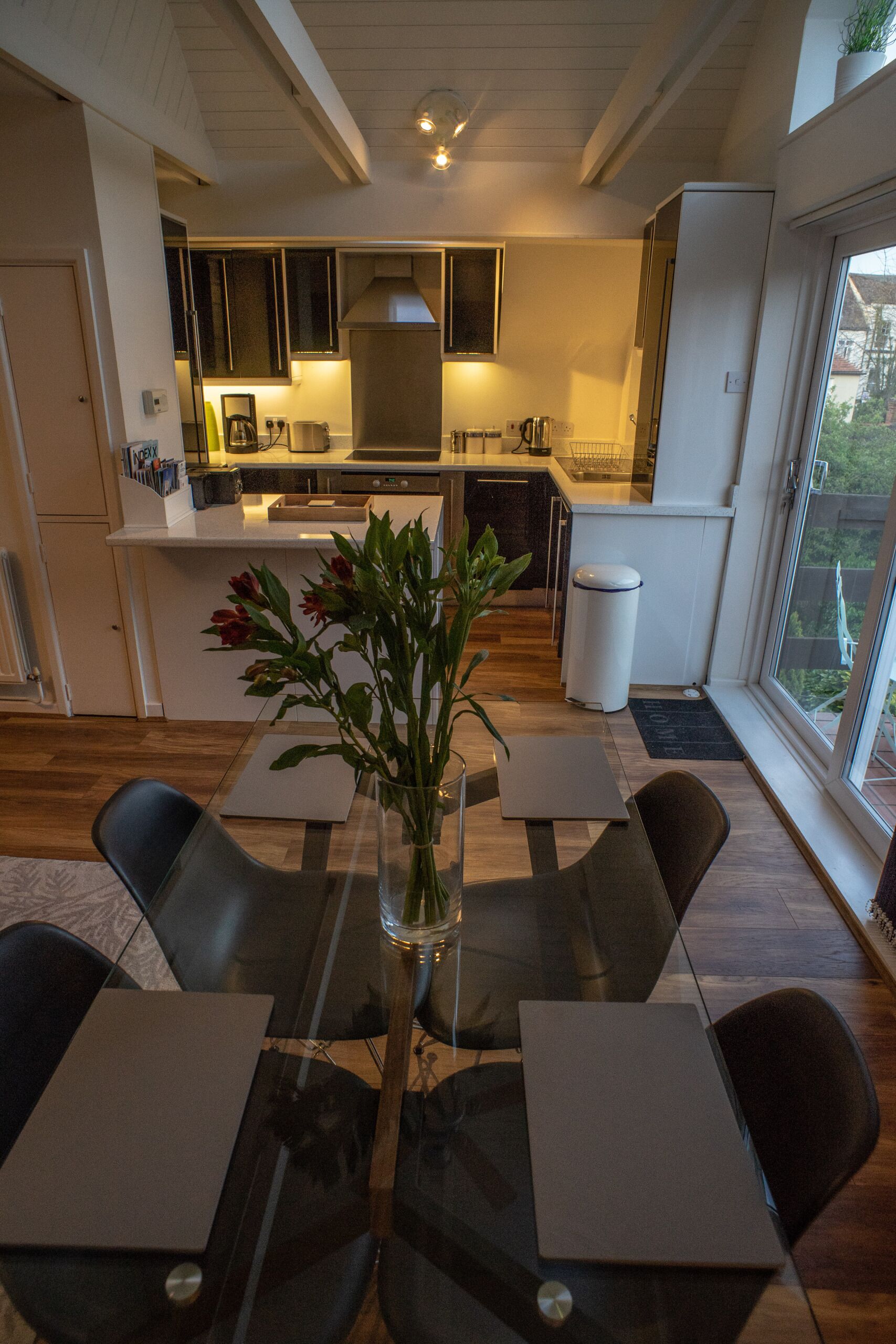 A modern kitchen with a central island, a dining area with a glass table, and a view of the outdoors through large windows.