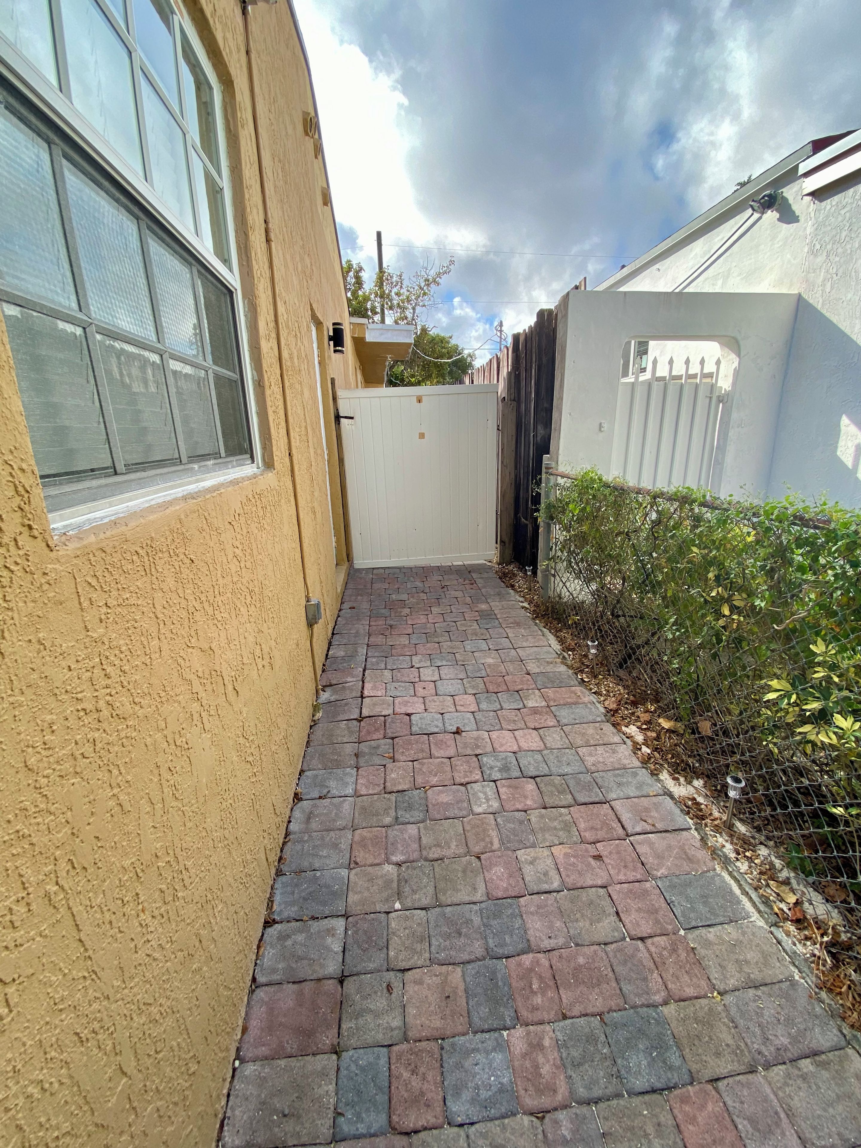 A narrow paved walkway leading to a gated area with a yellow building on one side and a white fence on the other.