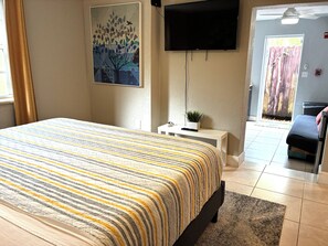A bedroom with a bed, a flat-screen TV, a painting, a small table with a plant, and a door leading to another room.