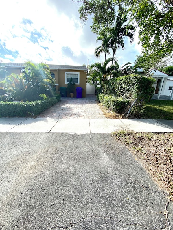 A paved driveway leading to a building with a window and a door, surrounded by greenery and a clear sky.