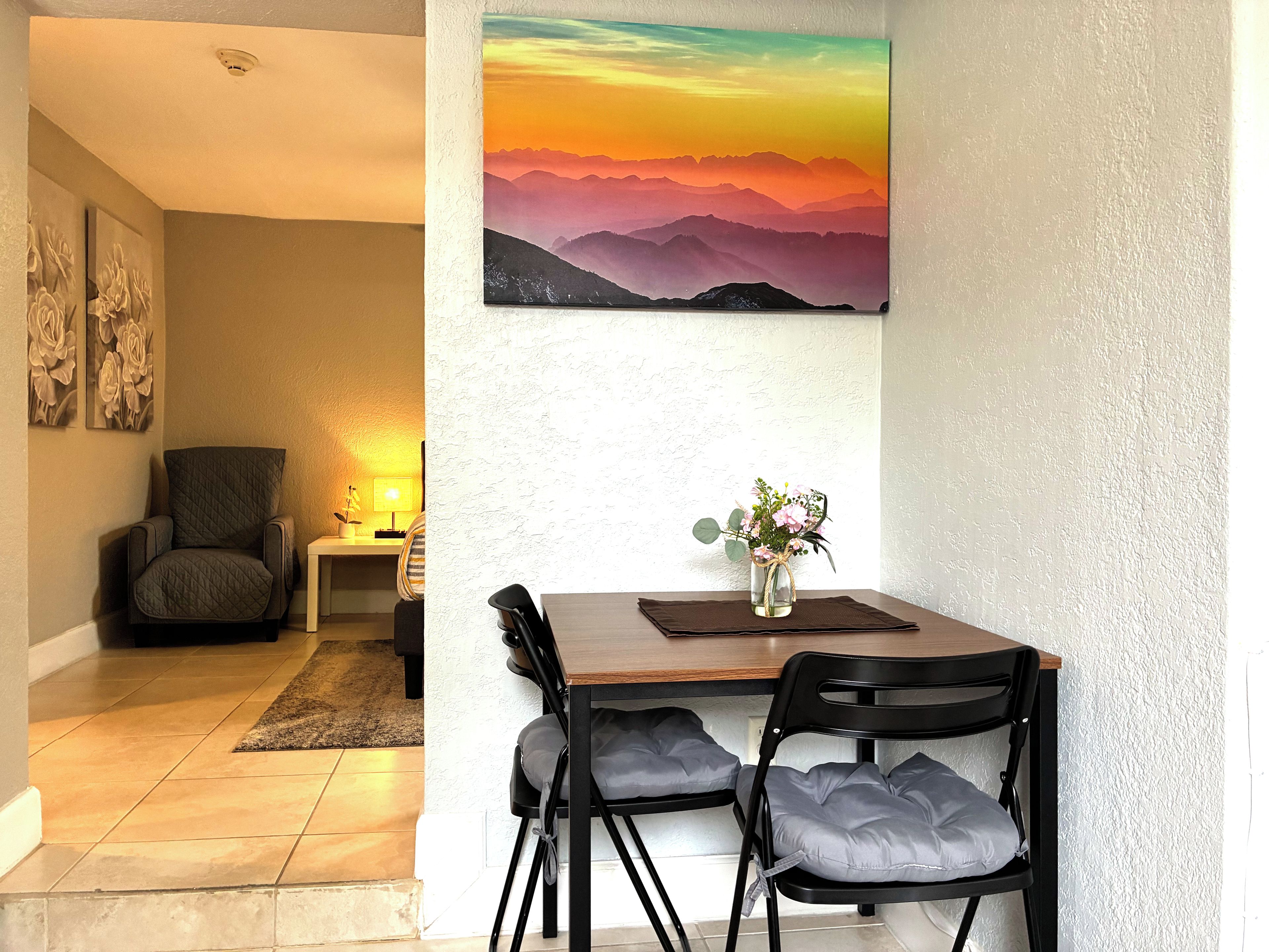 A dining area with a table, chairs, a vase with flowers, and a painting of a sunset.
