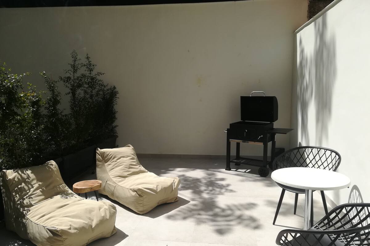 A patio with a beige sofa, a small round table, a grill, and two chairs.