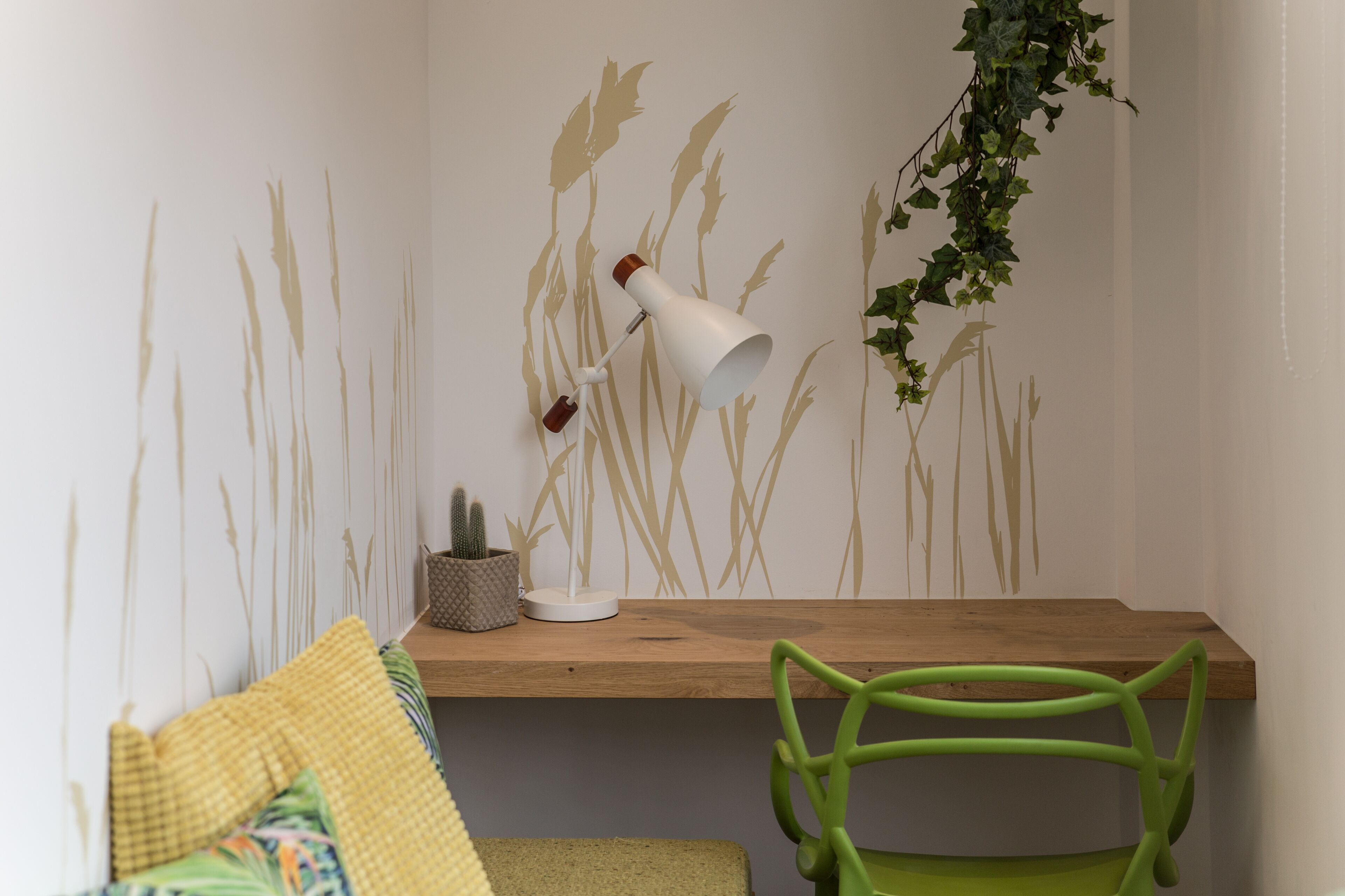 A room with a wooden desk, a green chair, a white lamp, and a wall with a plant and a wall decal.