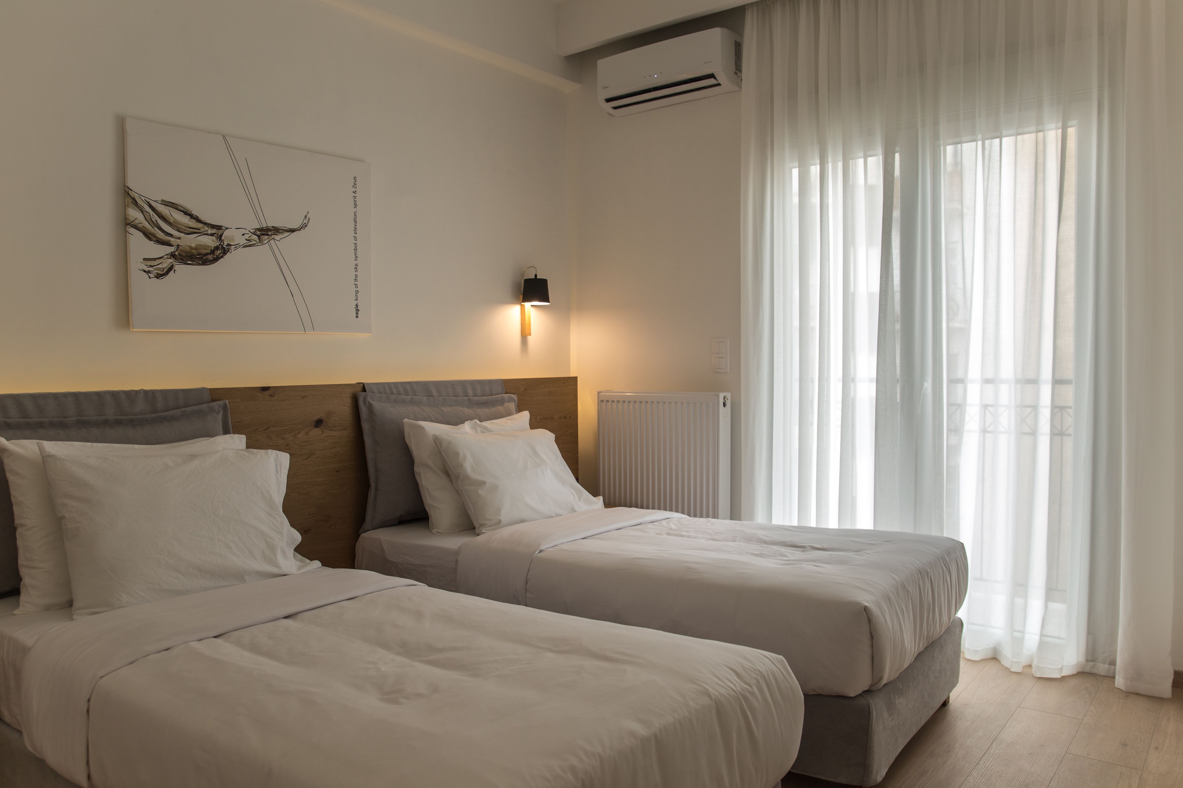 A hotel room with two beds, a wooden headboard, a wall-mounted artwork, a window with sheer curtains, and an air conditioning unit.