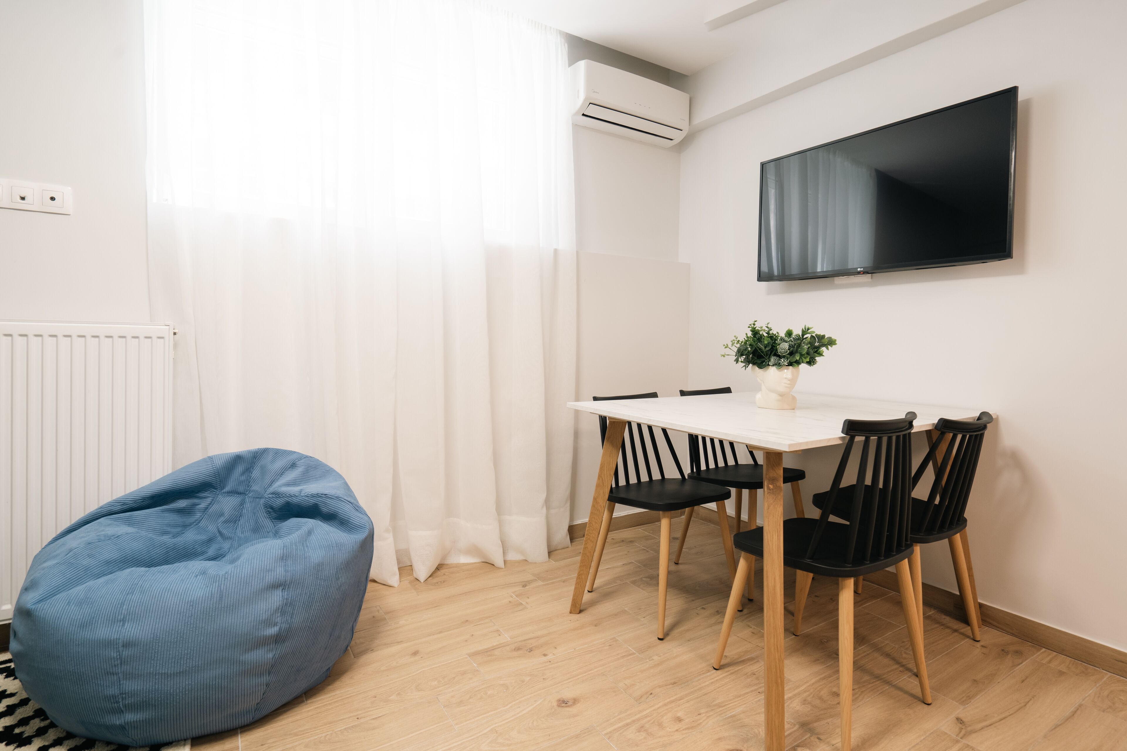 A modern room with a white table, four black chairs, a blue bean bag, a potted plant, and a flat-screen TV on the wall.