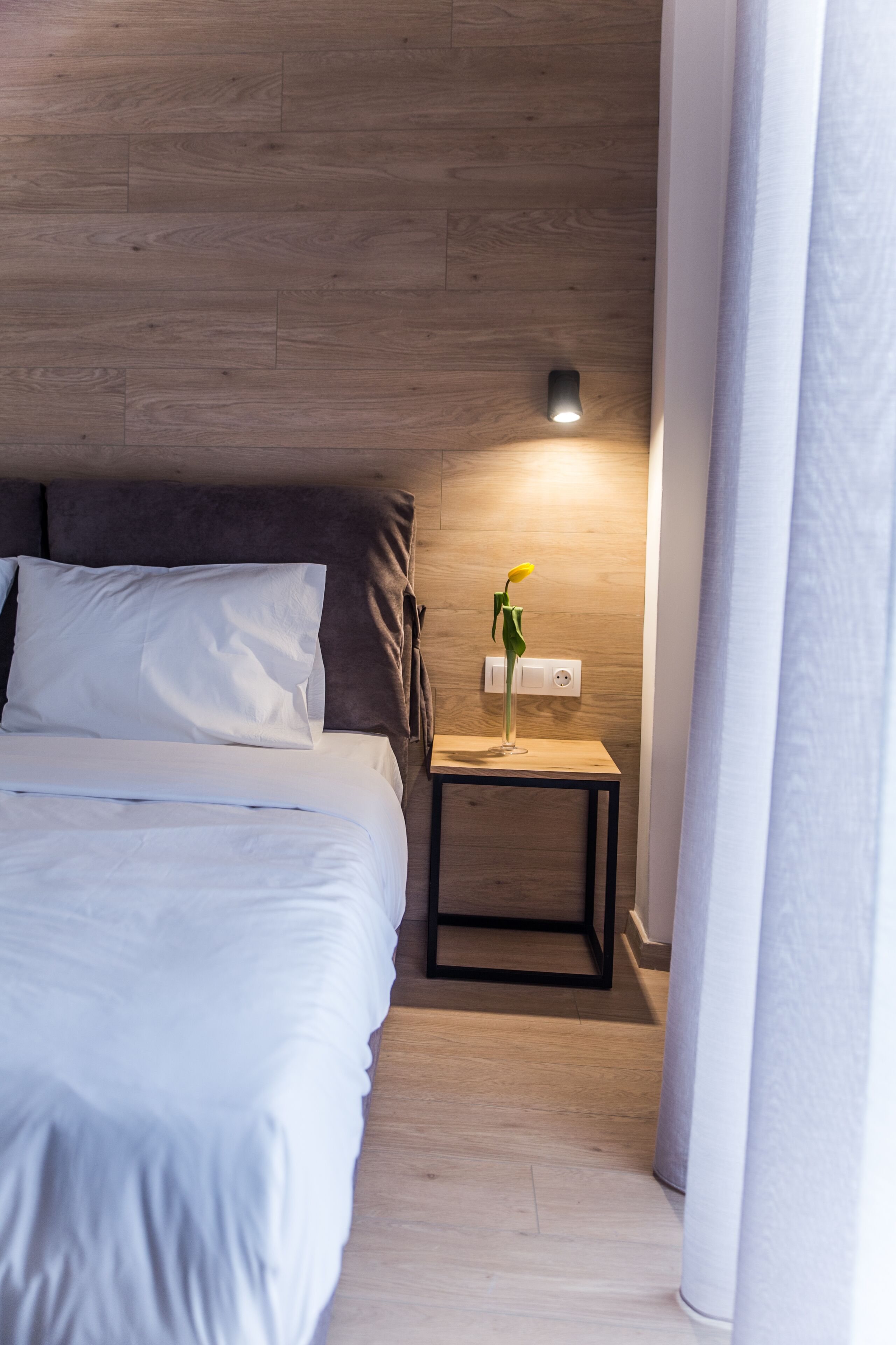 A neatly made bed with white linens, a small bedside table with a lamp, and a wall-mounted switch.