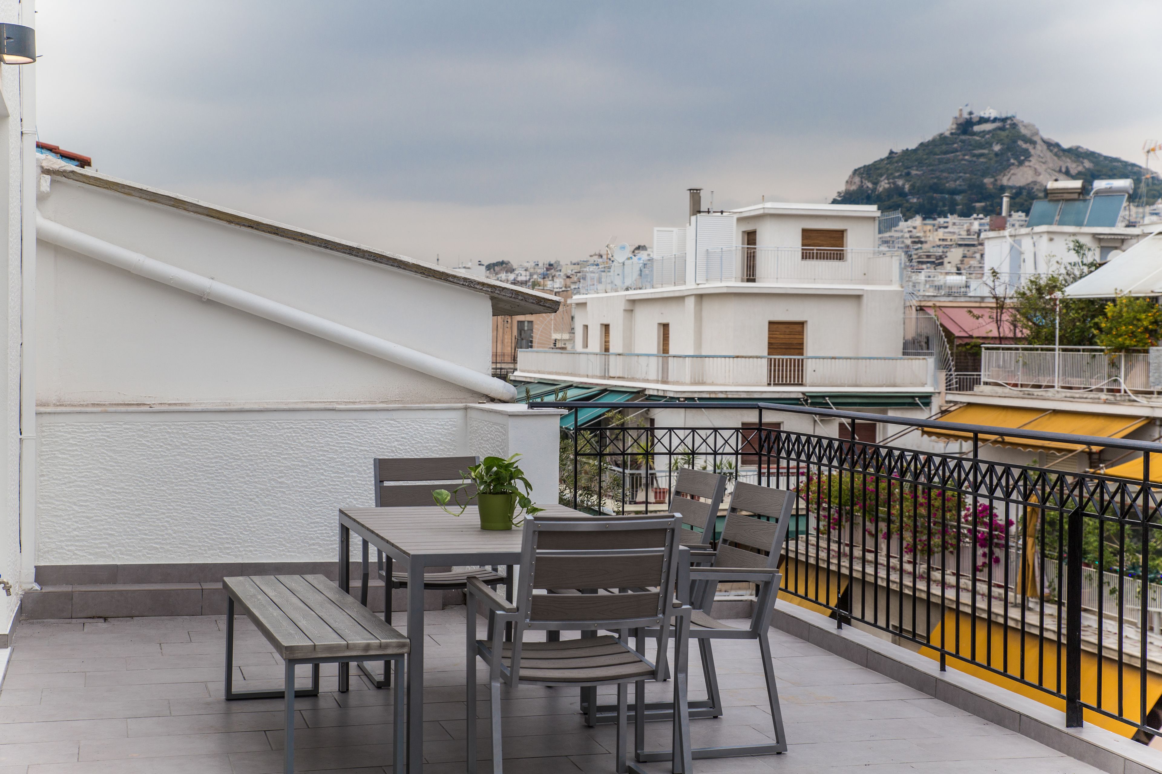 Une terrasse sur le toit, avec une table et des chaises, un pot de fleurs et une vue sur le paysage urbain, avec une colline bien visible à l’arrière-plan.