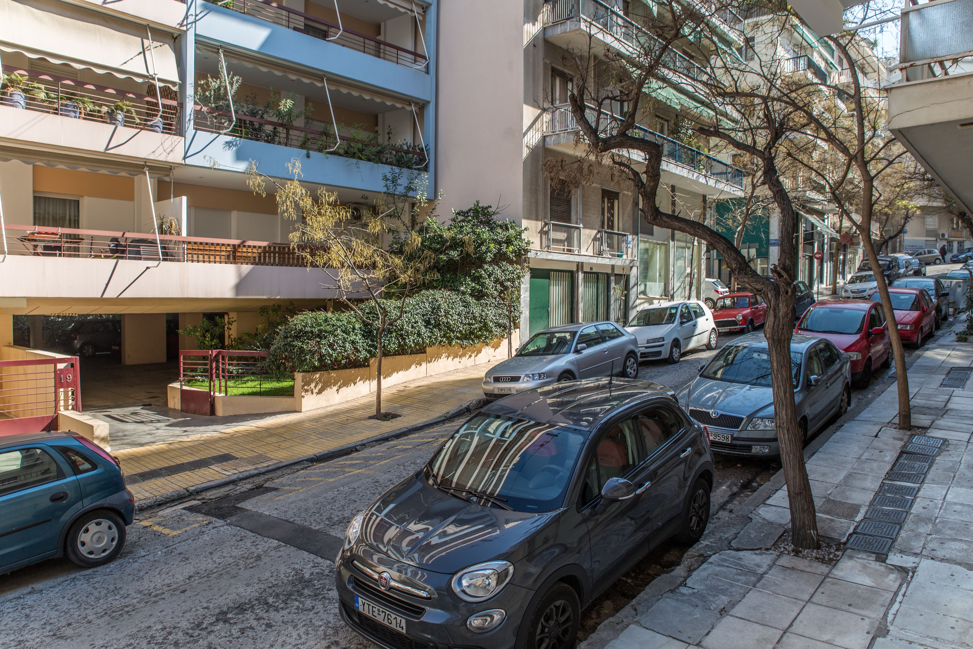 Un immeuble résidentiel à plusieurs étages, doté de balcons, avec des voitures garées et une rue bordée d’arbres.