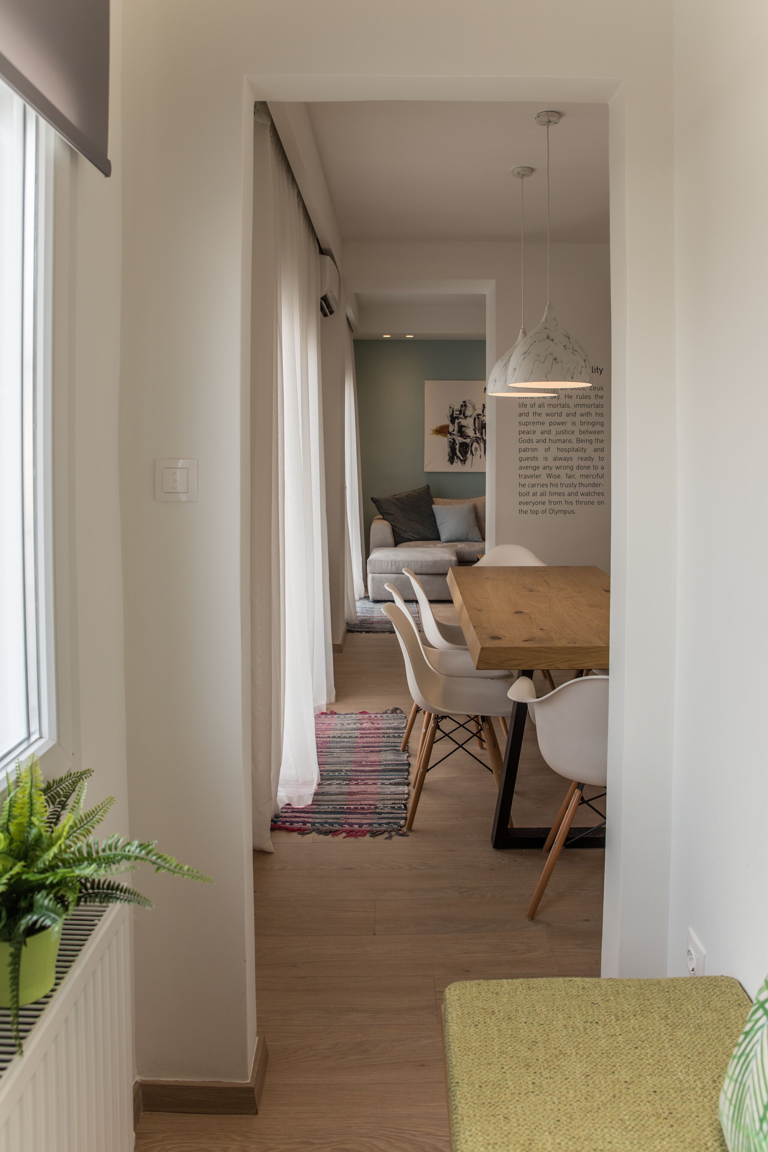 Un pasillo estrecho con un comedor que presenta una mesa de madera y sillas blancas, una planta en maceta y vistas a una sala de estar con un sofá y un cuadro.