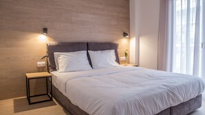 Un dormitorio moderno con cabecera de madera, una cama grande con ropa de cama blanca, dos mesitas de noche con lámparas y una ventana con cortinas translúcidas.