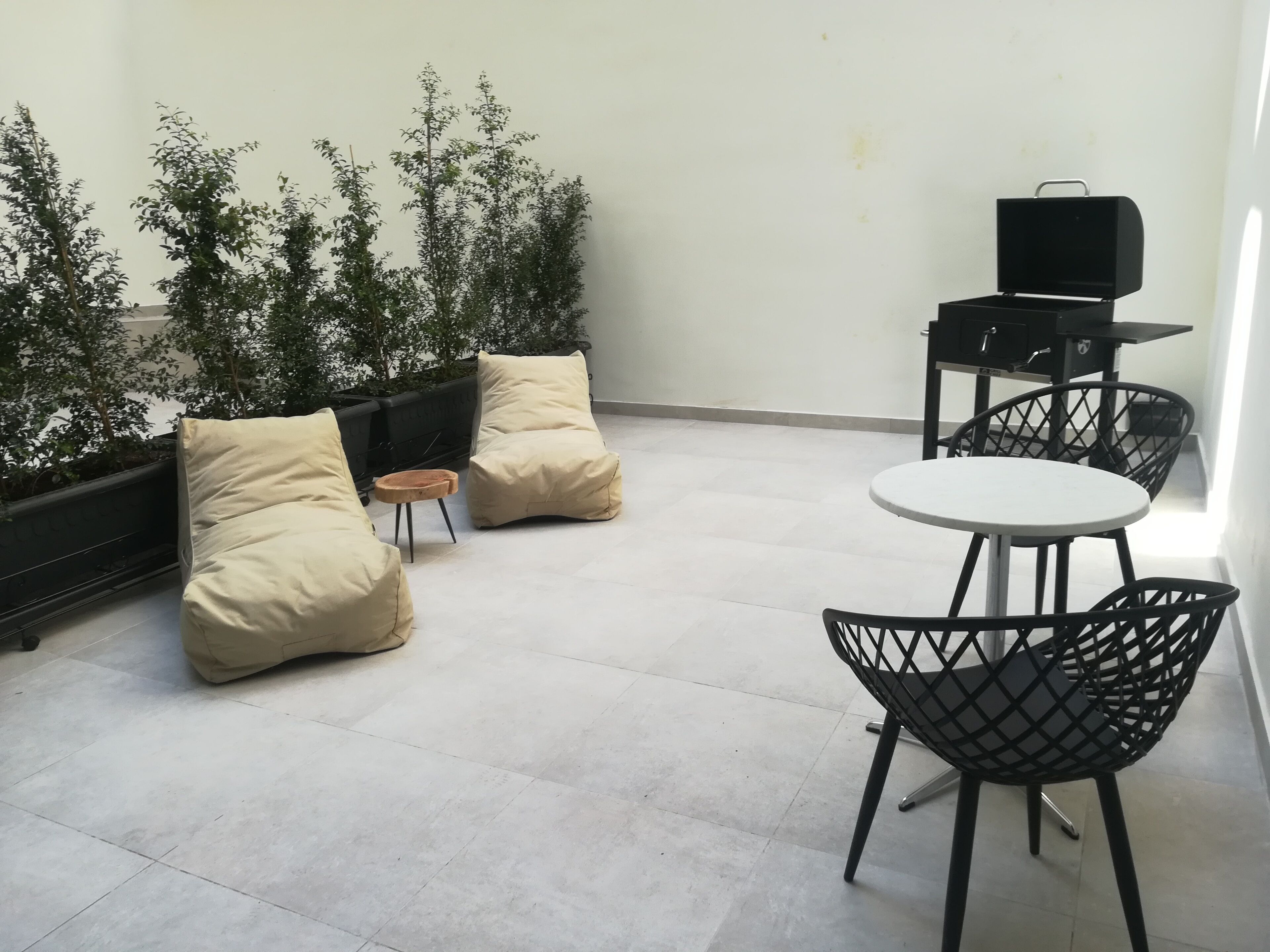 A rooftop terrace with two beige cushioned chairs, a small round table, a black grill, and a black chair with a crisscross design.
