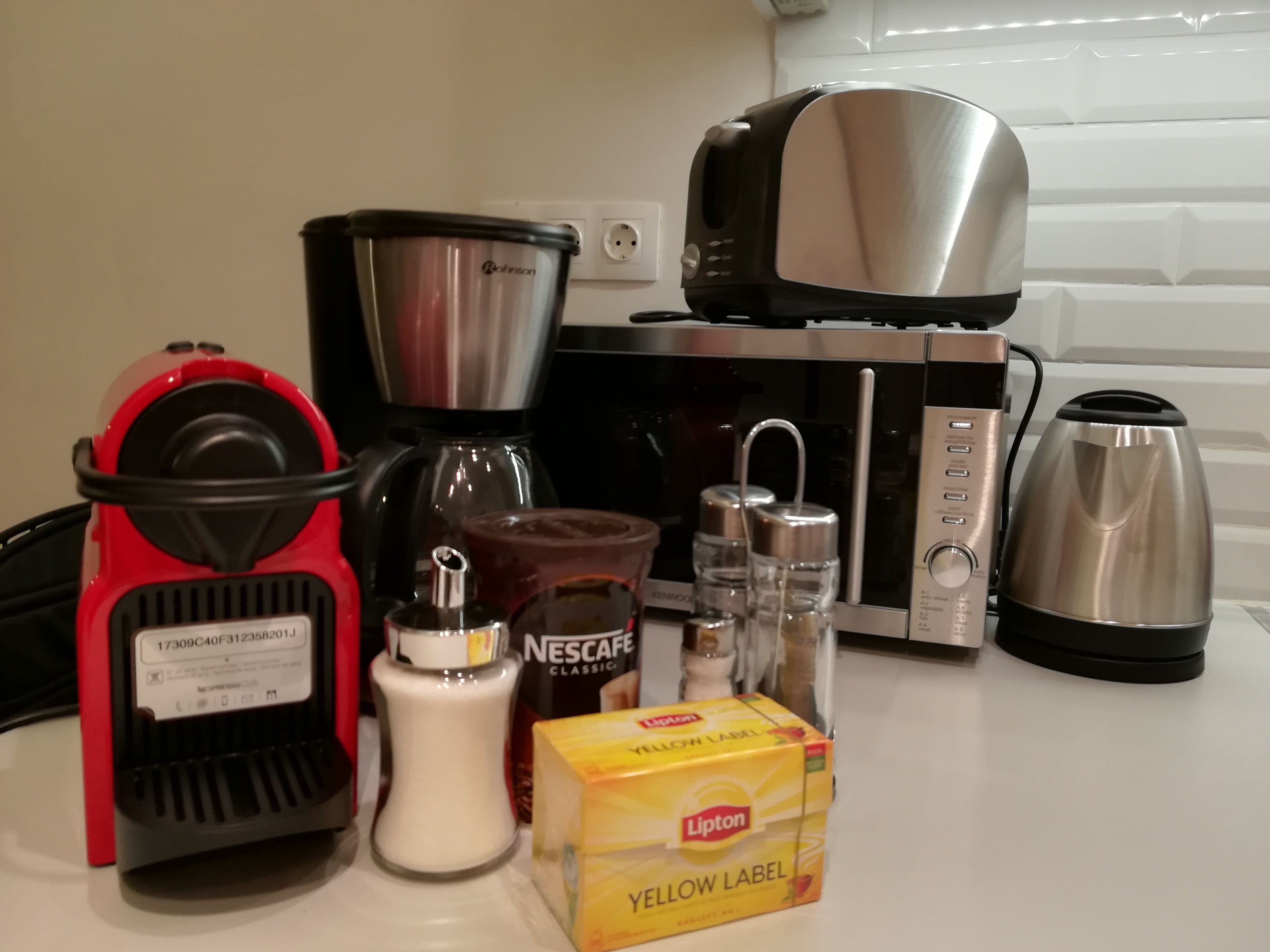 Uma cozinha com cafeteira, torradeira, chaleira, uma caixa de chá Lipton e uma cafeteira Nescafe.